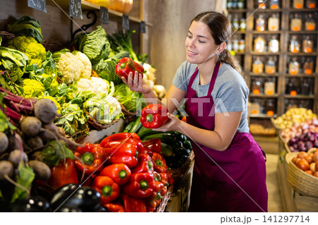 Female seller holding red bell pepper standing in fruit and vegetable section of supermarket Female seller holding red bell pepper standing in fruit and vegetable section of supermarket 141297714