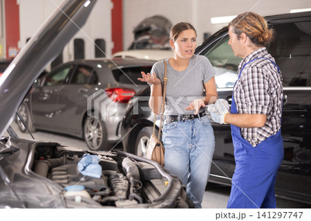 Woman talking to auto mechanic in uniform about repairing his car engine in auto repair shop Woman talking to auto mechanic in uniform about repairing his car engine in auto repair shop 141297747