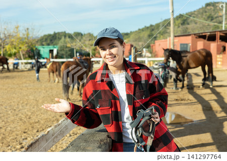 Portrait of positive young female stable worker holding horse halter or harness near wooden fence on ranch 141297764
