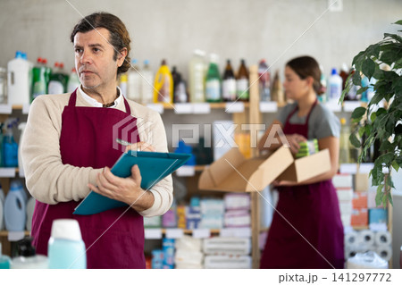 Man seller, with paper and invoice in hands, make inventory in store, girl employee in background Man seller, with paper and invoice in hands, make inventory in store, girl employee in background 141297772