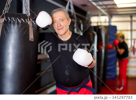 Senior karate practitioner resting against punching bag in gym Senior karate practitioner resting against punching bag in gym 141297783