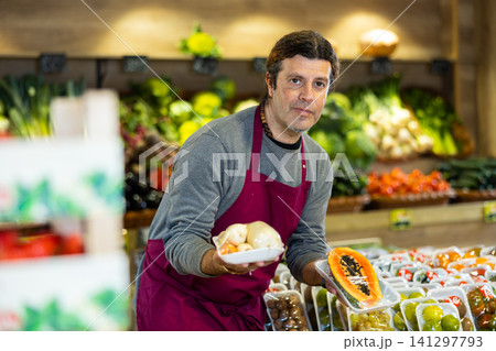 Male seller holding packaged carrots and sliced papaya standing in fruit and vegetable section of supermarket Male seller holding packaged carrots and sliced papaya standing in fruit and vegetable section of supermarket 141297793