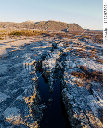 Aerial view of a tectonic fissure and frosty landscape in Thingvellir, Iceland, with distant mountains under golden morning light. 141297803