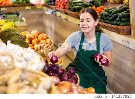 Female grocery store worker arranges red onion and other vegetables on counter and display case Female grocery store worker arranges red onion and other vegetables on counter and display case 141297850