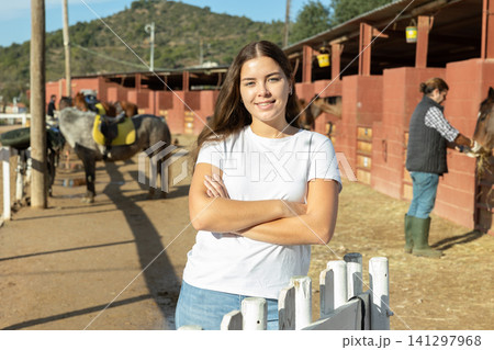 Portrait of young woman in front of stable and paddock for walking horses Portrait of young woman in front of stable and paddock for walking horses 141297968