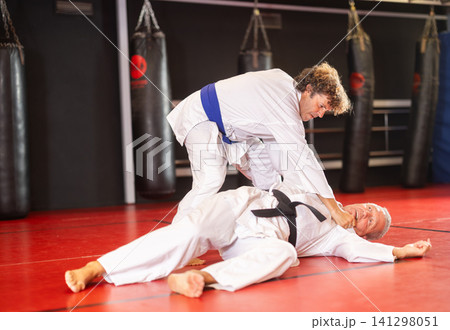 Focused man in white gi with blue belt practicing judo techniques during sparring with experienced black-belt opponent on red tatami in training room .. 141298051