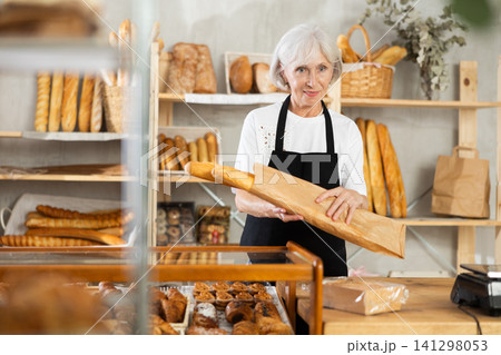 Mature saleswoman holding paper bag with baguettes in bakery Mature saleswoman holding paper bag with baguettes in bakery 141298053