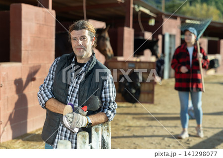 Adult male stables worker in plaid shirt holding brushes and gloves before cleaning horses from dirt in ranch Adult male stables worker in plaid shirt holding brushes and gloves before cleaning horses from dirt in ranch 141298077