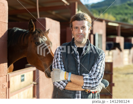 Adult male stables worker in plaid shirt holding brushes and gloves before cleaning horses from dirt in ranch Adult male stables worker in plaid shirt holding brushes and gloves before cleaning horses from dirt in ranch 141298124