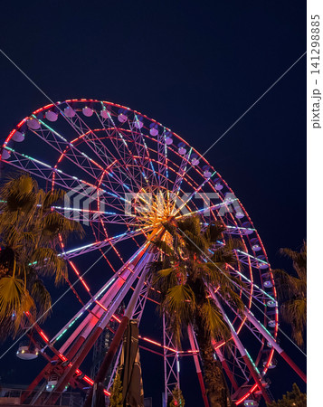 Illuminated Ferris wheel at night in Batumi, Georgia with colorful neon lights reflecting on palm leaves under dark sky Illuminated Ferris wheel at night in Batumi, Georgia with colorful neon lights reflecting on palm leaves under dark sky 141298885