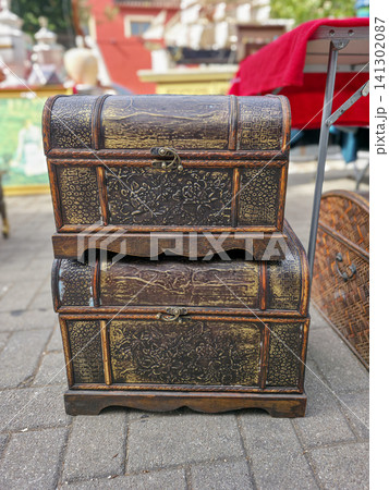 Two wooden treasure chests with gold patterns sit stacked at an outdoor flea market. The sun shines on them as people browse nearby stalls. 141302087