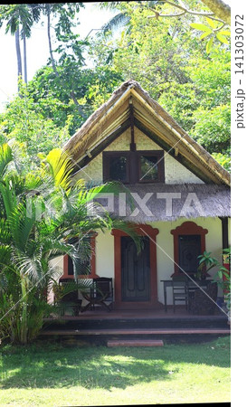 Thatched roof bungalow facade with woven bamboo gable, carved wooden doors, terracotta trim and palm shaded tropical garden in Bali resort setting for peaceful island stay 141303072