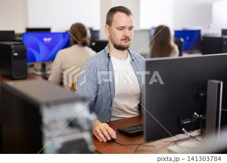 Guy student of computer courses looks at monitor screen, types on keyboard and does work 141303784