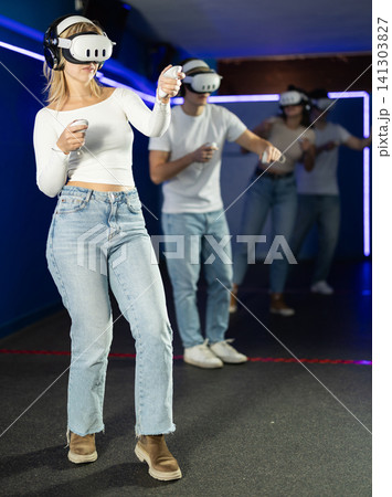 In darkened hall of gaming club, girl in VR helmet takes part in group game 141303827