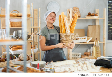 Bakery female seller stands proudly in front of fresh bread holding basket of freshly baked baguettes in kitchen 141303843