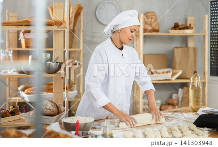 Young baker girl is standing near work table in kitchen, working with dough 141303948