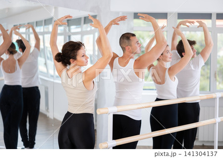 Rear view of young beautiful woman in white top and black leggings practicing classic ballet postures and movements at barre during group classes in mirrored choreography studio Rear view of young beautiful woman in white top and black leggings practicing classic ballet postures and movements at barre during group classes in mirrored choreography studio 141304137