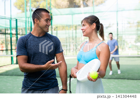 Portrait of two woman and man in sportswear on outdoor court with paddle racket and ball in hands after match, talking friendly Portrait of two woman and man in sportswear on outdoor court with paddle racket and ball in hands after match, talking friendly 141304448