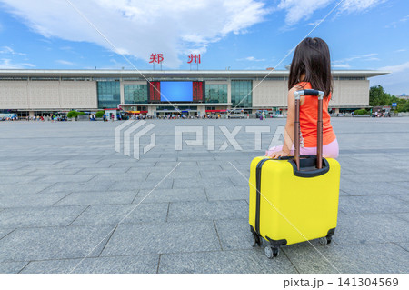 A tired little girl sits on her yellow suitcase in the station square, with modern buildings and a skyline in the background. Above the station are  large characters: "zhengzhou." 141304569