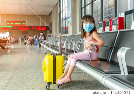 Young Asian Schoolgirl Wearing a Blue Medical Face Mask Sitting in a Waiting Hall of a Train Station for Security During a Journey. Young Asian Schoolgirl Wearing a Blue Medical Face Mask Sitting in a Waiting Hall of a Train Station for Security During a Journey. 141304571