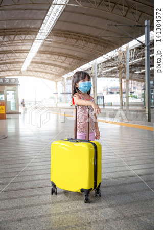 Vertical Photo of an Asian Kid Girl Wearing a Medical Face Mask Standing on a Train Station Platform with a Yellow Luggage. Vertical Photo of an Asian Kid Girl Wearing a Medical Face Mask Standing on a Train Station Platform with a Yellow Luggage. 141304572