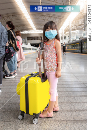 Vertical Photo of an Asian Kid Girl Wearing a Medical Face Mask Standing on a Train Station Platform with a Yellow Luggage.The text above reads: "Convenient Transfers" and "Exit". Vertical Photo of an Asian Kid Girl Wearing a Medical Face Mask Standing on a Train Station Platform with a Yellow Luggage.The text above reads: "Convenient Transfers" and "Exit". 141304573