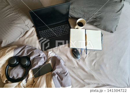 Laptop, notebook, coffee and headphones scattered on an unmade bed. Concept of remote work blurring boundaries with rest and burnout Laptop, notebook, coffee and headphones scattered on an unmade bed. Concept of remote work blurring boundaries with rest and burnout 141304722