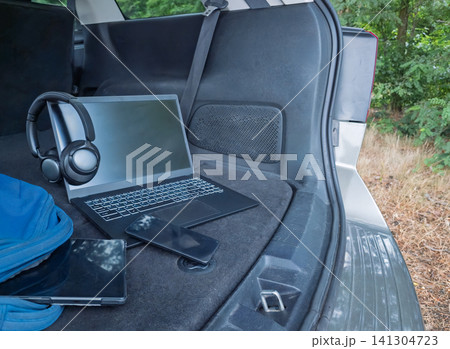 A laptop, smartphone, tablet and headphones are arranged in an open car trunk facing the forest. Concept of digital nomad workspace, remote work, and mobile office. A laptop, smartphone, tablet and headphones are arranged in an open car trunk facing the forest. Concept of digital nomad workspace, remote work, and mobile office. 141304723