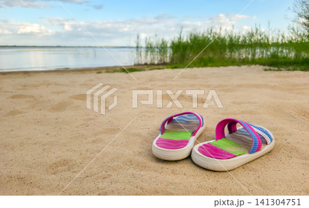 Bright colorful sandals rest in the sand on a lake beach with reeds beyond. Concept of summer vacation, leisure, and beach lifestyle with copy space. Bright colorful sandals rest in the sand on a lake beach with reeds beyond. Concept of summer vacation, leisure, and beach lifestyle with copy space. 141304751