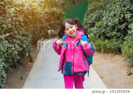 Full shot of a smiling school girl with pigtails wearing a pink down jacket and blue jeans, carrying a blue backpack, walking along a paved garden path with green shrubs. Full shot of a smiling school girl with pigtails wearing a pink down jacket and blue jeans, carrying a blue backpack, walking along a paved garden path with green shrubs. 141305839