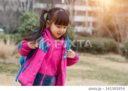 Close up three-quarter view of a smiling young girl with pigtails wearing a pink down jacket and a blue backpack, looking back over her shoulder with a blurred garden in warm sunlight. Close up three-quarter view of a smiling young girl with pigtails wearing a pink down jacket and a blue backpack, looking back over her shoulder with a blurred garden in warm sunlight. 141305854