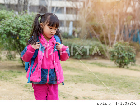 Close up three-quarter view of a smiling young girl with pigtails wearing a pink down jacket and a blue backpack, looking back over her shoulder with a blurred garden in warm sunlight. 141305856