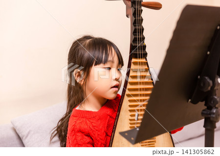 Young Chinese girl practicing Pipa traditional musical instrument on a sofa at home, creative arts and music education. Young Chinese girl practicing Pipa traditional musical instrument on a sofa at home, creative arts and music education. 141305862