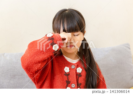 Young East Asian elementary girl rubbing itchy eye with one fist while eyes are closed, standing indoors against wall with gray sofa. Young East Asian elementary girl rubbing itchy eye with one fist while eyes are closed, standing indoors against wall with gray sofa. 141306014
