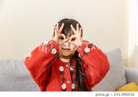 Playful East Asian preschool-age girl standing inside and pretending to form a telescope with both hands making circle gestures near her eyes Playful East Asian preschool-age girl standing inside and pretending to form a telescope with both hands making circle gestures near her eyes 141306016