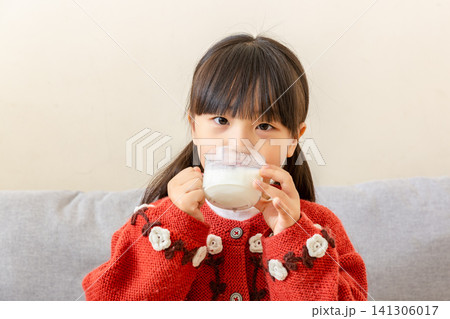 Cute Asian child in a red cardigan raising a clear glass cup to her mouth to take a sip of milk 141306017