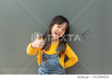 Happy Asian elementary girl standing against a green chalkboard and flashing a victory sign, wearing a yellow t-shirt and denim overalls Happy Asian elementary girl standing against a green chalkboard and flashing a victory sign, wearing a yellow t-shirt and denim overalls 141306052