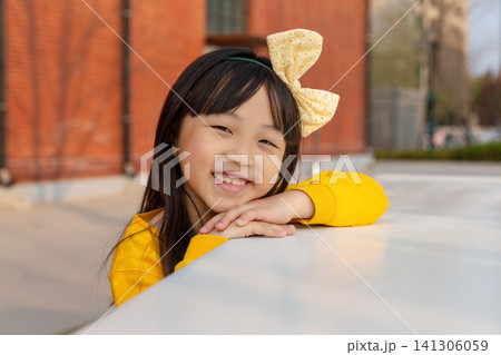 Happy young girl with pigtails and a yellow bow leaning comfortably against a white table and smiling broadly at the camera Happy young girl with pigtails and a yellow bow leaning comfortably against a white table and smiling broadly at the camera 141306059
