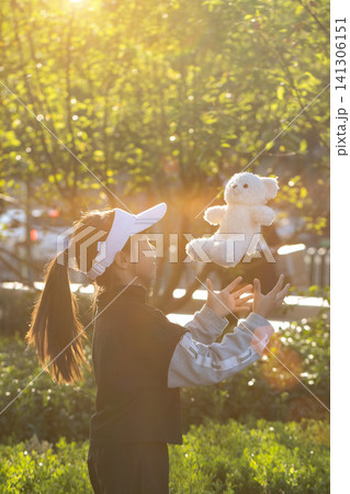 Cheerful little girl playing with teddy bear in park at sunset with golden sunlight 141306151