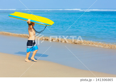Portrait of little surfer boy with bodyboard have fun on beach, walk by white sand beach. Summer adventures, active families, parents and kids lifestyle, sports activities on school holiday with kids Portrait of little surfer boy with bodyboard have fun on beach, walk by white sand beach. Summer adventures, active families, parents and kids lifestyle, sports activities on school holiday with kids 141306865