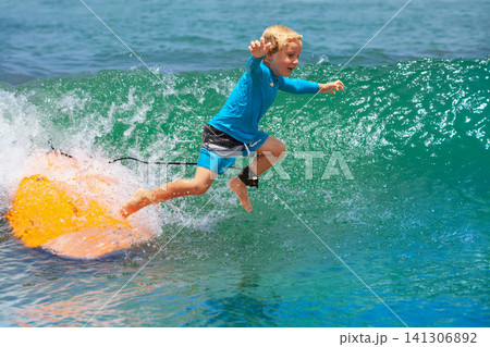 Happy young surfer boy with surfboard have fun on beach, riding on small ocean waves. Summer adventures, active families, parents and kids lifestyle, sports activities on school holiday with kids Happy young surfer boy with surfboard have fun on beach, riding on small ocean waves. Summer adventures, active families, parents and kids lifestyle, sports activities on school holiday with kids 141306892