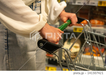 Close-up of Hands Gripping Shopping Cart Handle in Grocery Store Aisle, Focused on the Act of Shopping for Everyday Essentials and Groceries 141307421