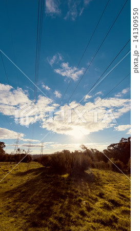 Low angle perspective of high voltage power lines cutting through a grassy countryside landscape against a bright sunset sky 141309550