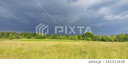 Dark storm clouds over green trees and rural grass field landscape Dark storm clouds over green trees and rural grass field landscape 141311416