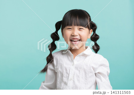 A happy East Asian schoolgirl laughing joyfully  while standing against a solid mint green studio background. 141311503