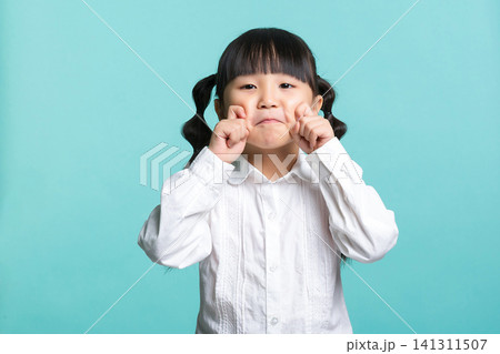 A playful elementary school girl pulling her cheeks with both hands to make a funny face against a light green background. 141311507