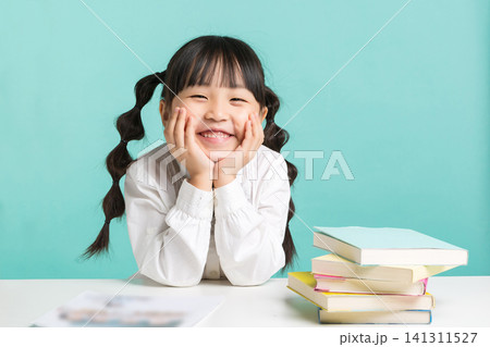 A sweet little schoolgirl leaning her chin on both hands while sitting at her study table with colorful books. A sweet little schoolgirl leaning her chin on both hands while sitting at her study table with colorful books. 141311527