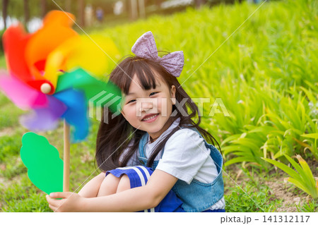 A charming young girl sits on a grassy slope with a bright rainbow pinwheel, smiling warmly at the camera with lush foliage behind. A charming young girl sits on a grassy slope with a bright rainbow pinwheel, smiling warmly at the camera with lush foliage behind. 141312117