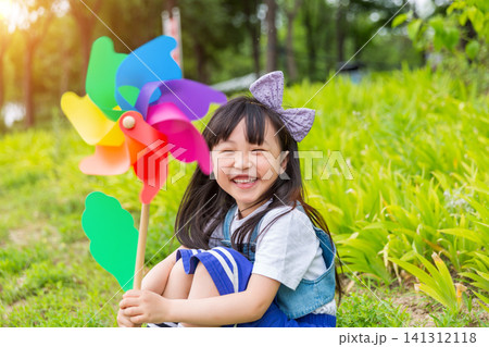 A charming young girl sits on a grassy slope with a bright rainbow pinwheel, smiling warmly at the camera with lush foliage behind. A charming young girl sits on a grassy slope with a bright rainbow pinwheel, smiling warmly at the camera with lush foliage behind. 141312118