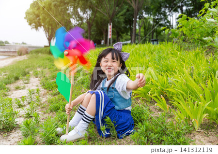 A charming young girl sits on a grassy slope with a bright rainbow pinwheel, smiling warmly at the camera with lush foliage behind. A charming young girl sits on a grassy slope with a bright rainbow pinwheel, smiling warmly at the camera with lush foliage behind. 141312119